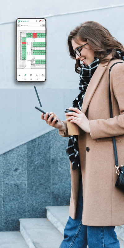 Desk booking system - how it can be used to benefit workplaces. Woman walking down stairs with a coffee in one hand, and a phone in the other. We can see a pop-up animation of the phone screen. She is making a booking for her desk using the Ronspot app.