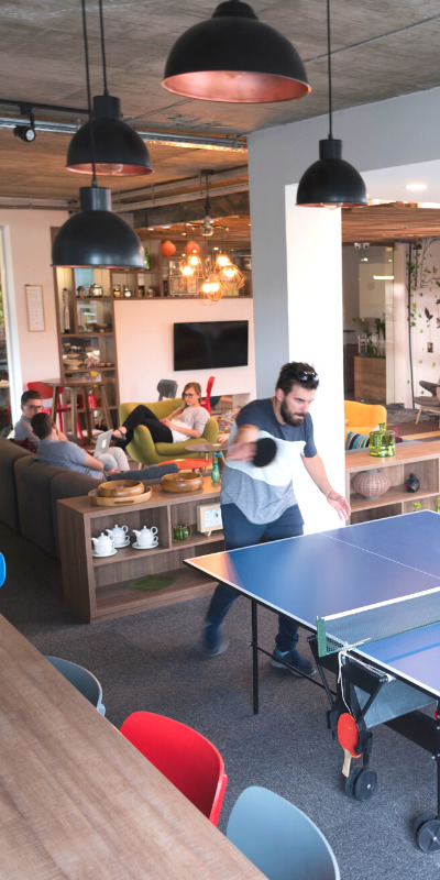 Workplace experience app - Workers enjoying a game of ping-pong in the office. Some co-workers are seen in the background sitting on couches speaking with each other